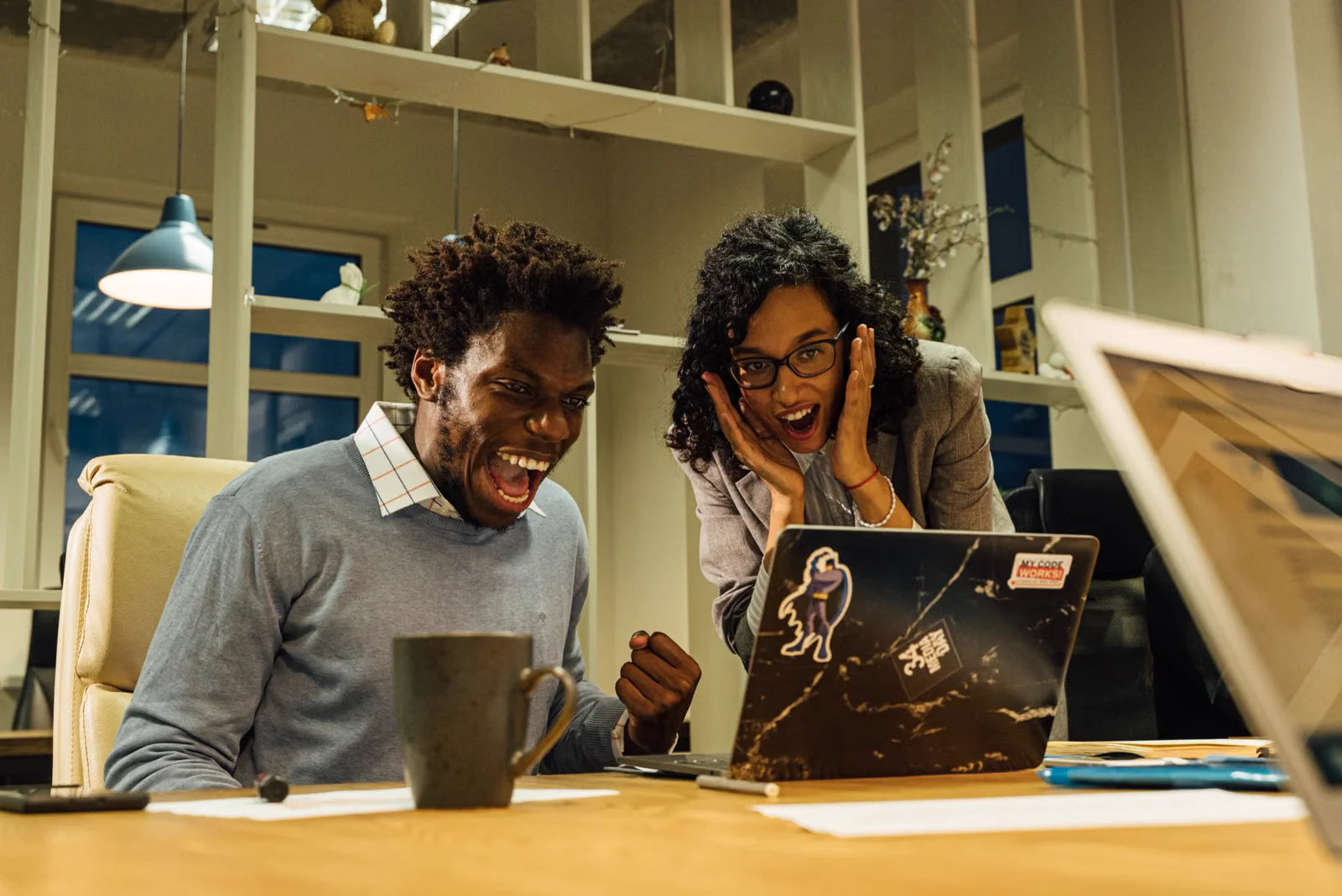 Two diverse colleagues laughing and cheering together at a warm office desk with a laptop